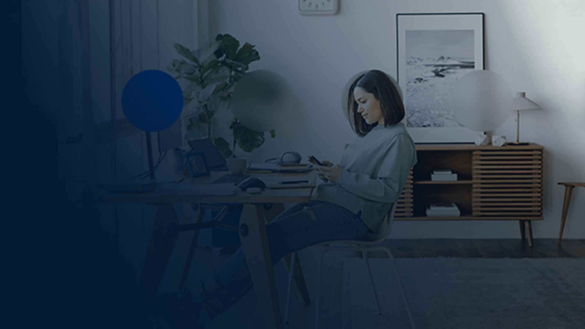A woman sat at a desk in her home office, reviewing her finances on a smart phone. This helps illustrate how Telefonica Tech helped boost Virgin Money's customer acquisition.