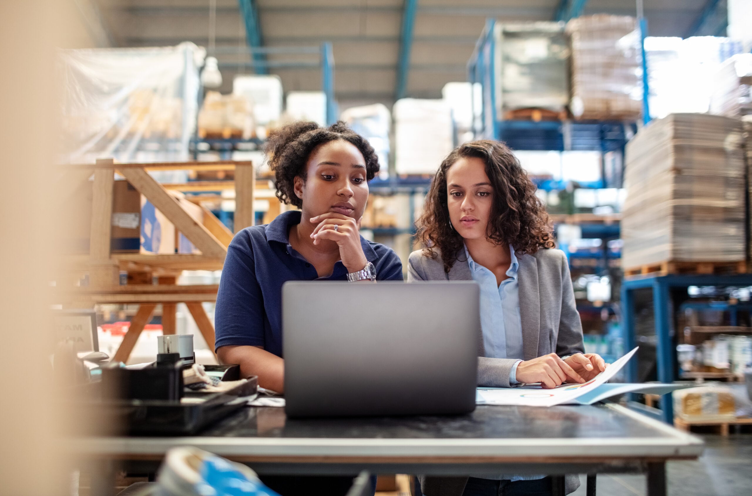 Two workers in a warehouse look at a laptop screen and some printed documents