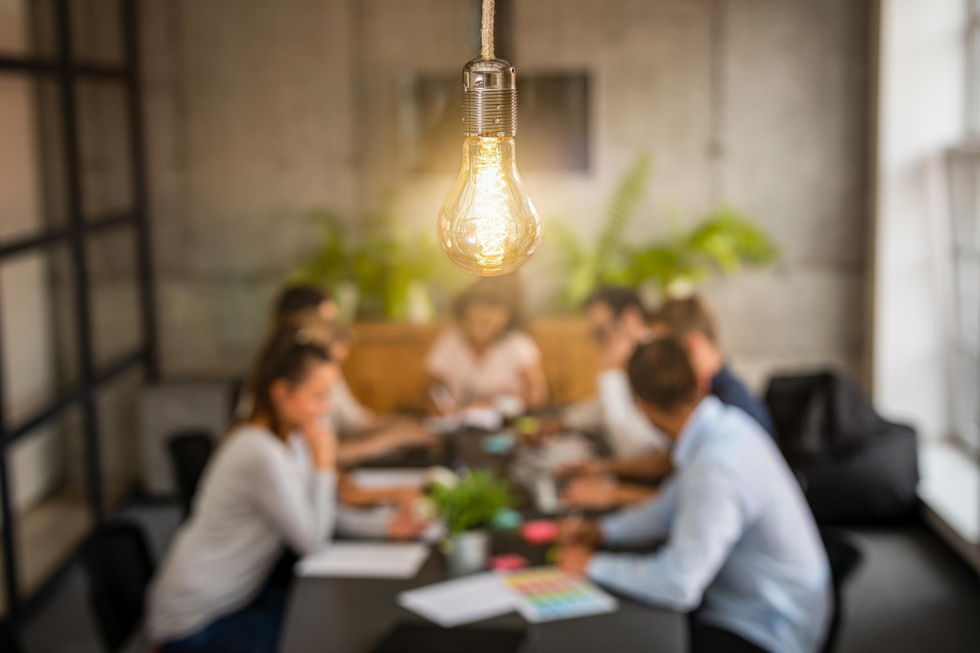 Primer plano de una bombilla iluminada y de fondo una mesa de trabajadores reunidos - Foreground of an illuminated light bulb and in the background a table of workers gathered together