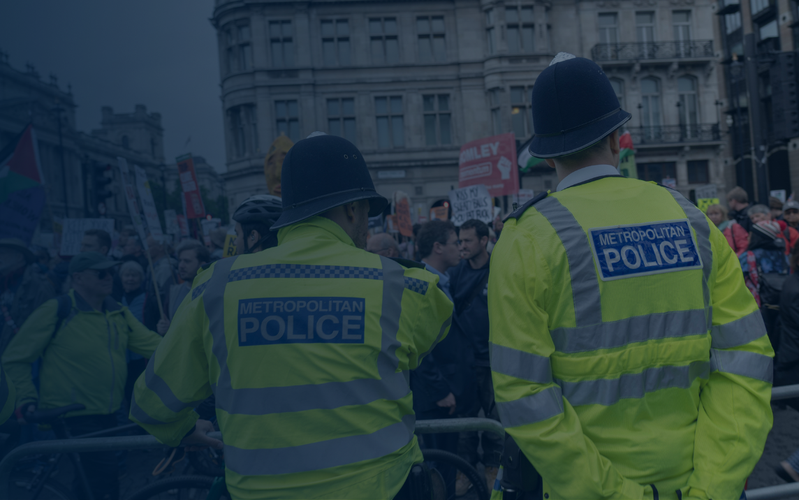 Metropolitan Police officers in high-visibility uniforms observe a large public demonstration from behind barriers, reflecting public order policing, proportionality, and legitimacy as measured by the PEEL assessment framework.
