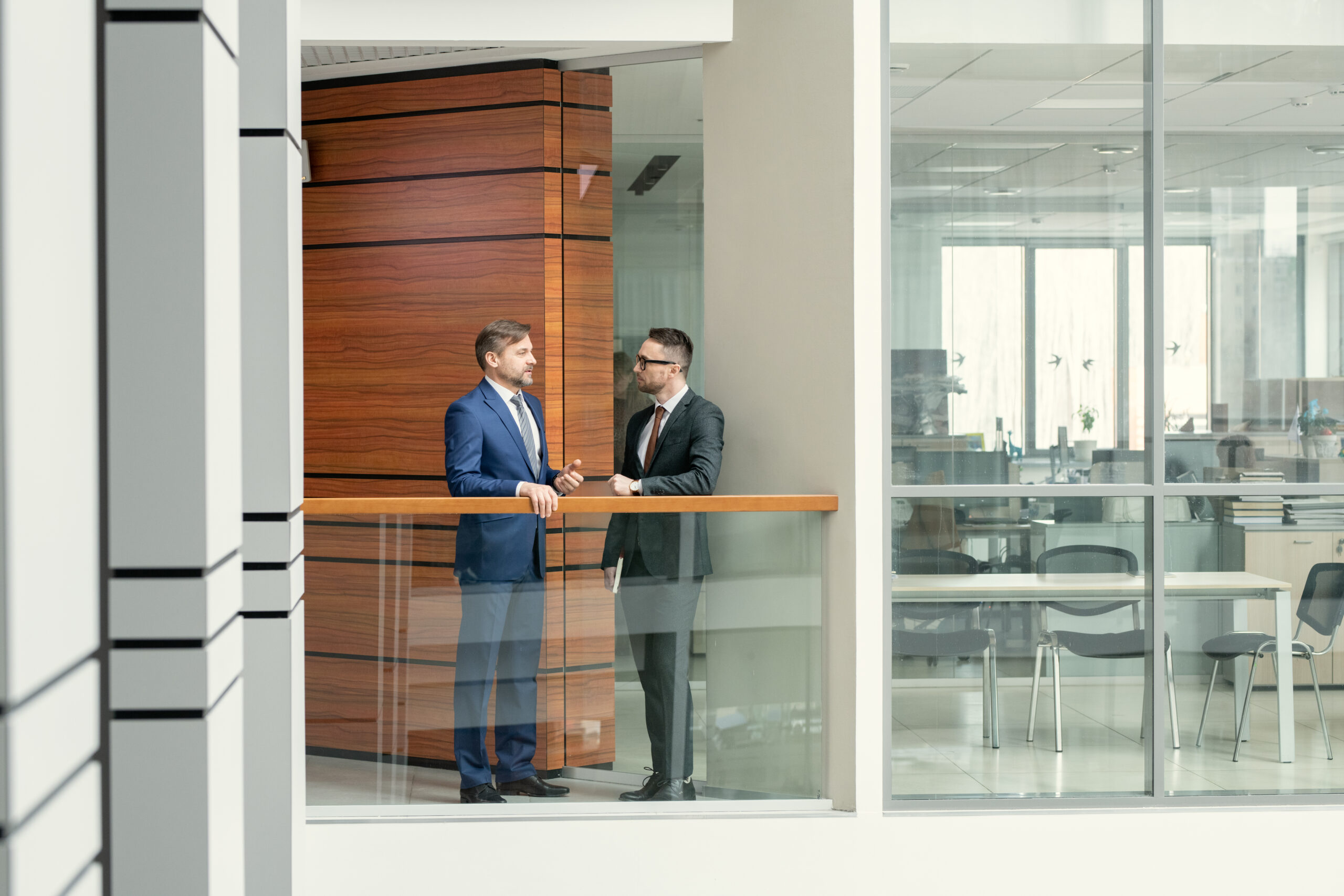 Mature businessman standing at railing and talking to young colleague in corridor