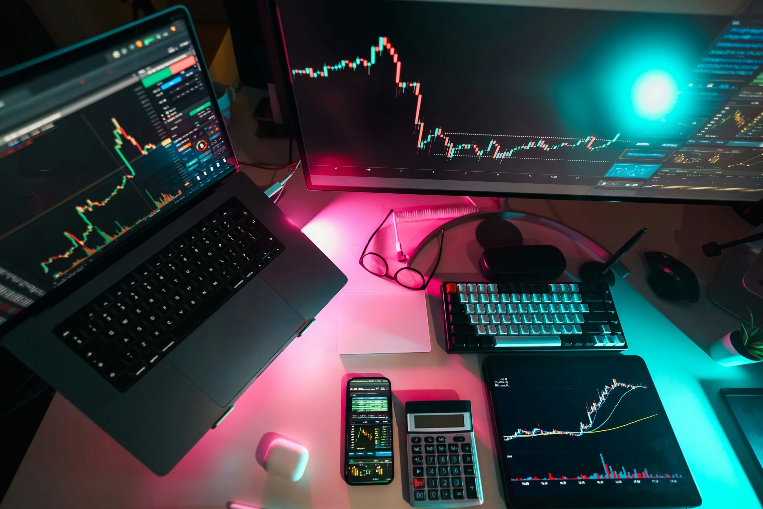 A modern trading workstation with a laptop, large external monitor, tablet, and smartphone displaying stock market candlestick charts. A mechanical keyboard, calculator, glasses, wireless earbuds, and mouse are arranged on a desk illuminated by teal and pink ambient lighting, creating a high-tech financial trading setup.